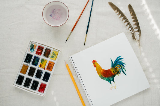 A notebook with a chicken art and near it is a watercolor and brush